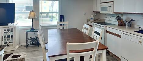 A modern kitchen with white cabinets, a wooden dining table, and white chairs. There is a TV, a lamp, and a window with blinds.