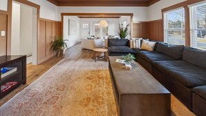 A spacious living room with a large sofa, a coffee table, and a rug. There is a ceiling fan and a window with blinds.