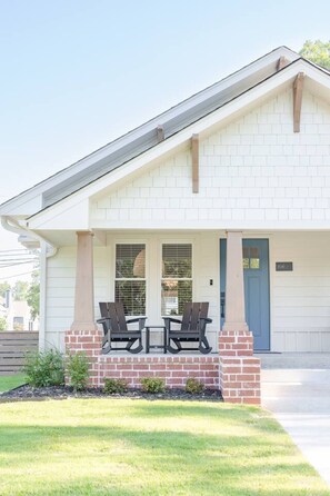 A two-story building with a white exterior, a blue door, and a covered porch with two chairs. - Luxury 2BR Home | Walk to Rose District | Sleeps 6 (Broken Arrow)