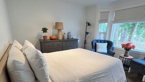 Ein modernes Schlafzimmer mit einem großen Bett, einem Schminktisch, einem Sessel und einem Fenster mit Blick auf Bäume.