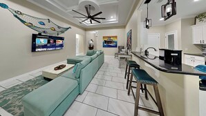 A modern living room with a sectional sofa, a TV mounted on the wall, a ceiling fan, and a kitchen area with a bar stool. - Ferryman's Landing by Miss Kitty’s (Aransas Pass)