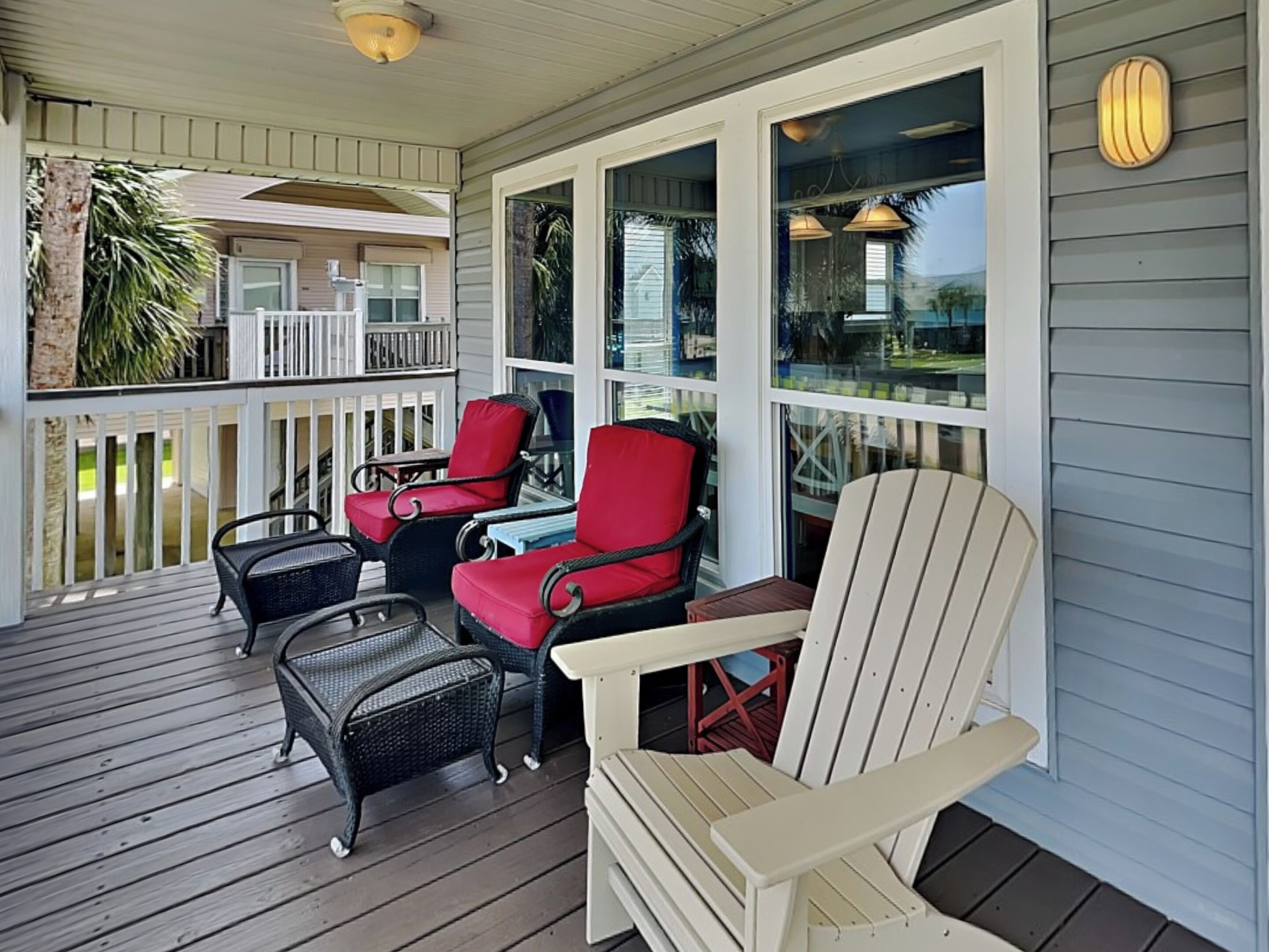 A covered porch with wicker chairs and a white Adirondack chair, wooden deck, and a view of the ocean through large windows.