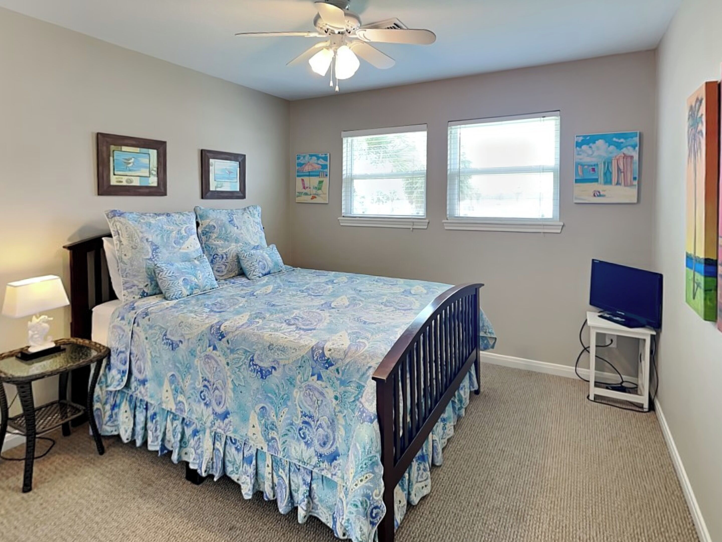 A bedroom with a bed, a ceiling fan, two windows, and artwork on the walls.