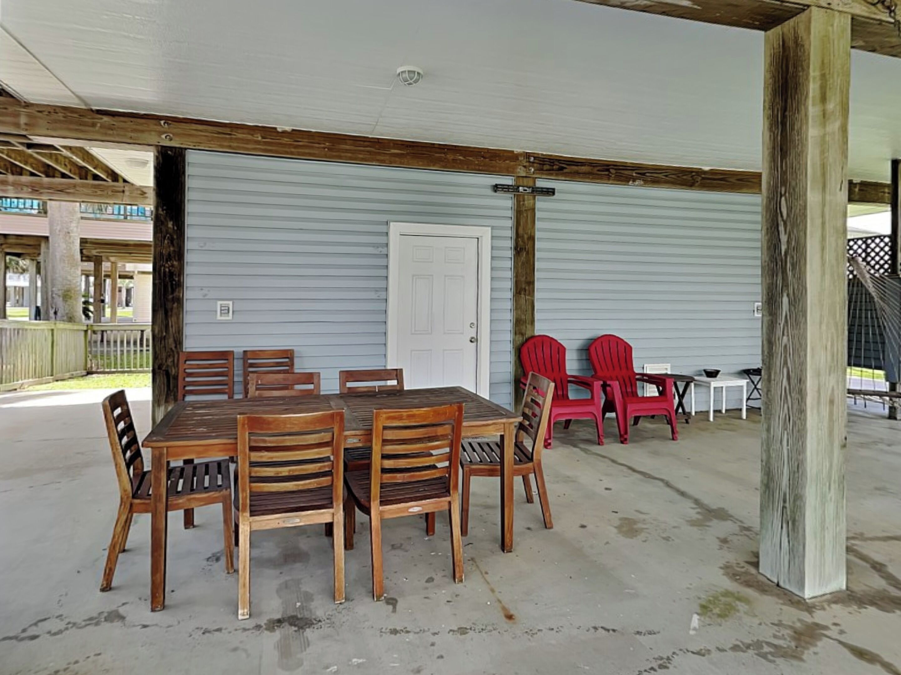 Un espace de restauration extérieur abrité, avec une table et des chaises en bois, une porte blanche et des chaises rouges en arrière-plan.