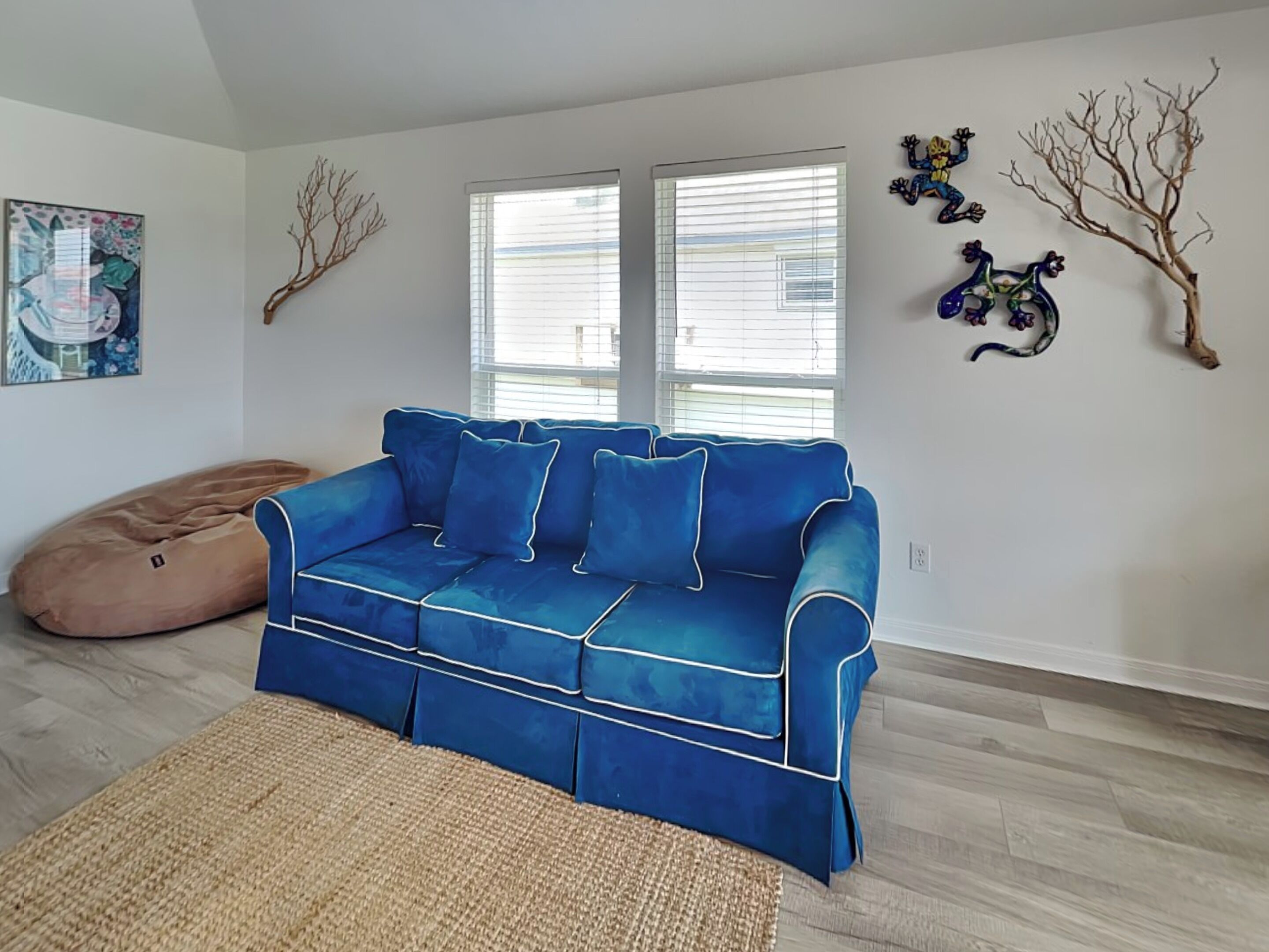Een woonkamer met een blauwe bank, een relaxfauteuil en een geweven vloerkleed. Twee ramen met jaloezieën, een schilderij en wanddecoratie.