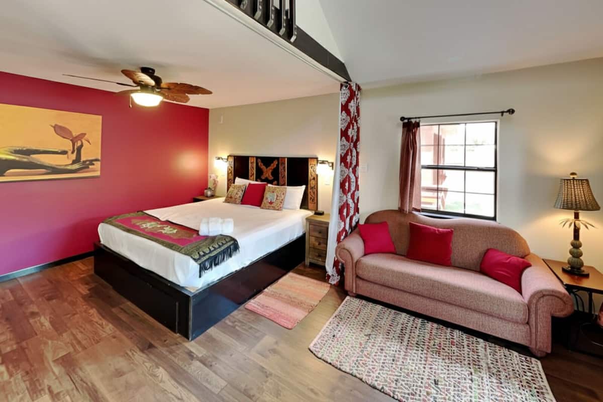A bedroom with a bed, a sofa, a window, a ceiling fan, and a painting on the wall.