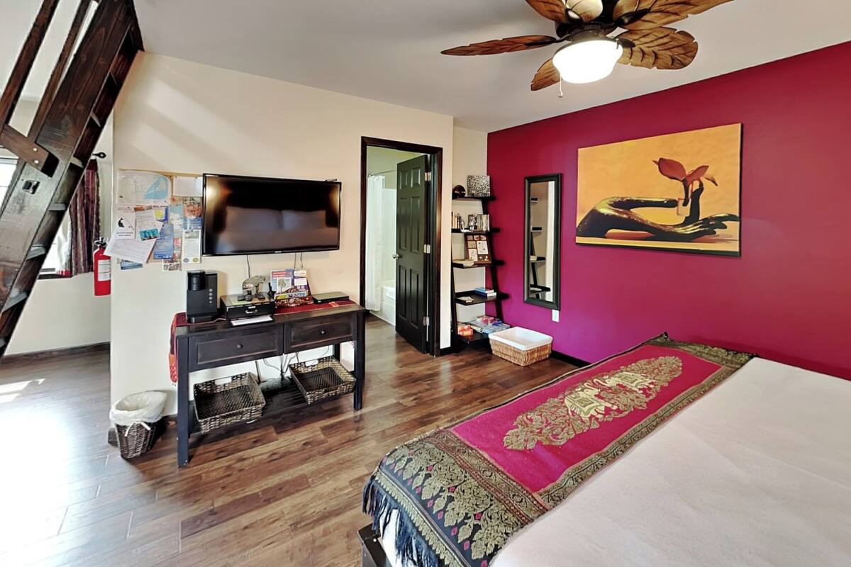 A bedroom with a bed, a desk with a TV, a ceiling fan, a painting, and a red wall.