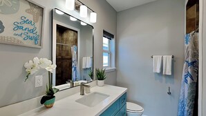 A bathroom with a white sink, blue cabinets, a large mirror, and a shower curtain with a blue and white pattern. - Bayside Abode by Miss Kitty’s (Rockport)