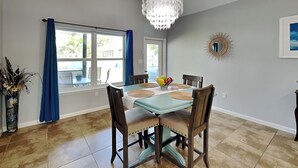 A dining area with a table set for six, a chandelier, and a view of the outdoors through large windows. - Bayside Abode by Miss Kitty’s (Rockport)