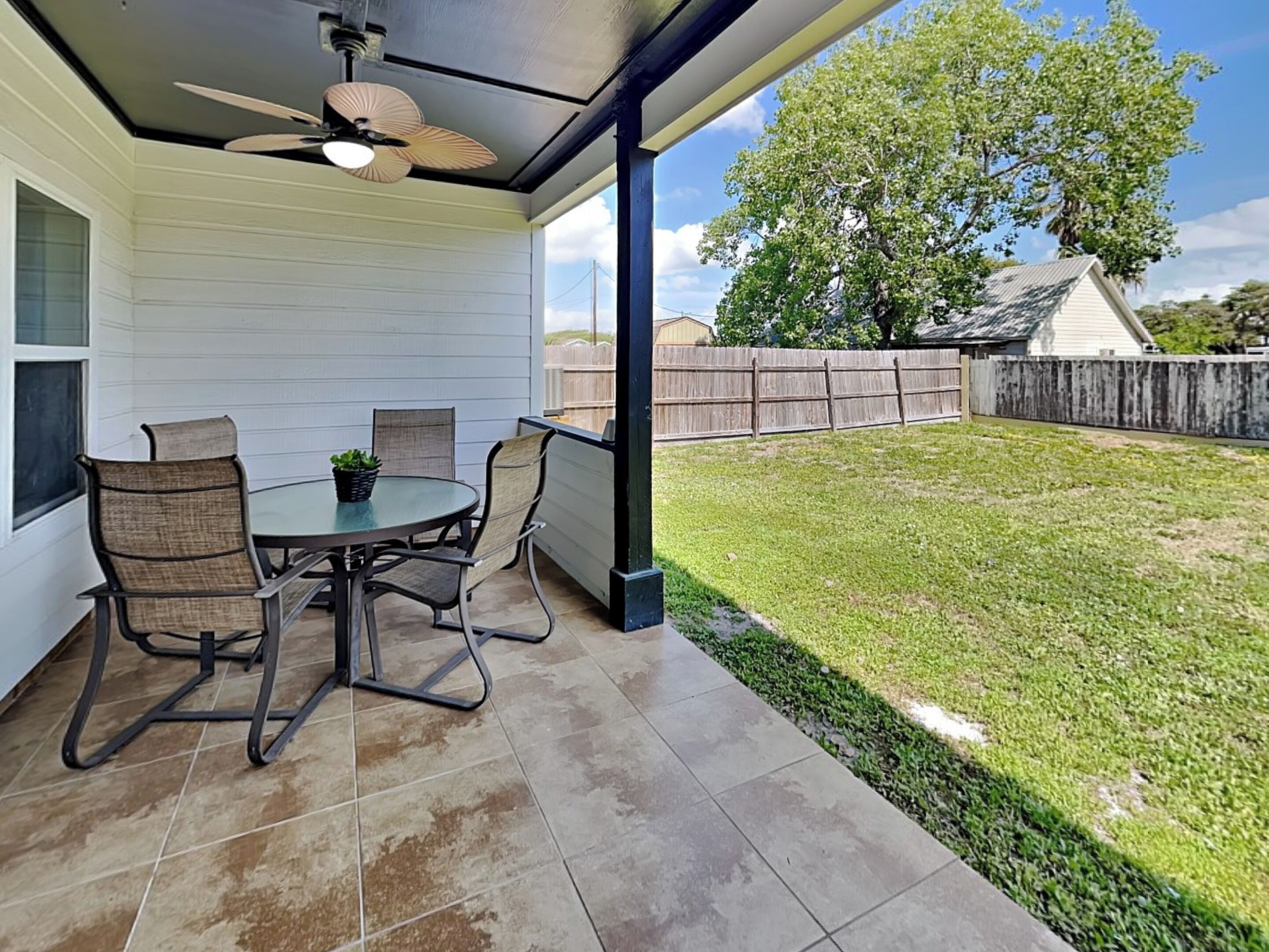 A covered patio with a table and chairs, a ceiling fan, and a view of a grassy yard with a wooden fence.