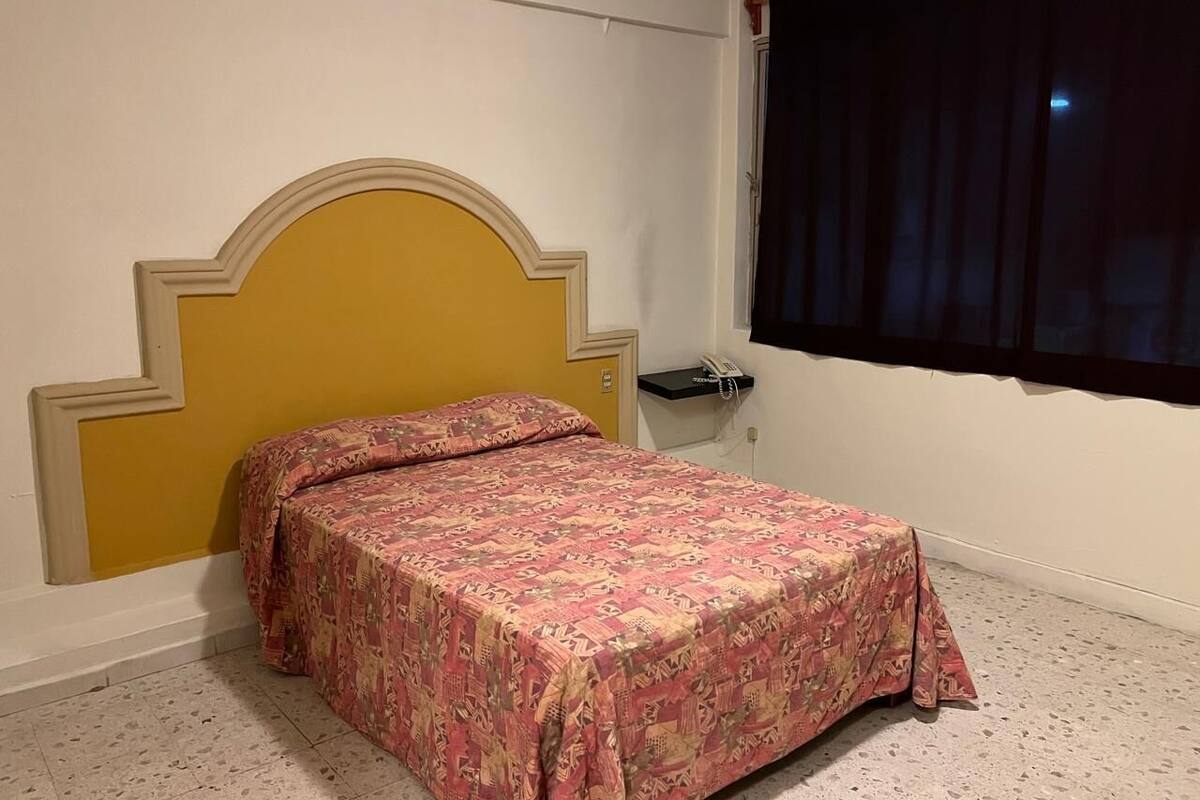 Ein Schlafzimmer mit Bett, Kopfstütze, Nachttisch, Telefon und einem Fenster mit Vorhängen.