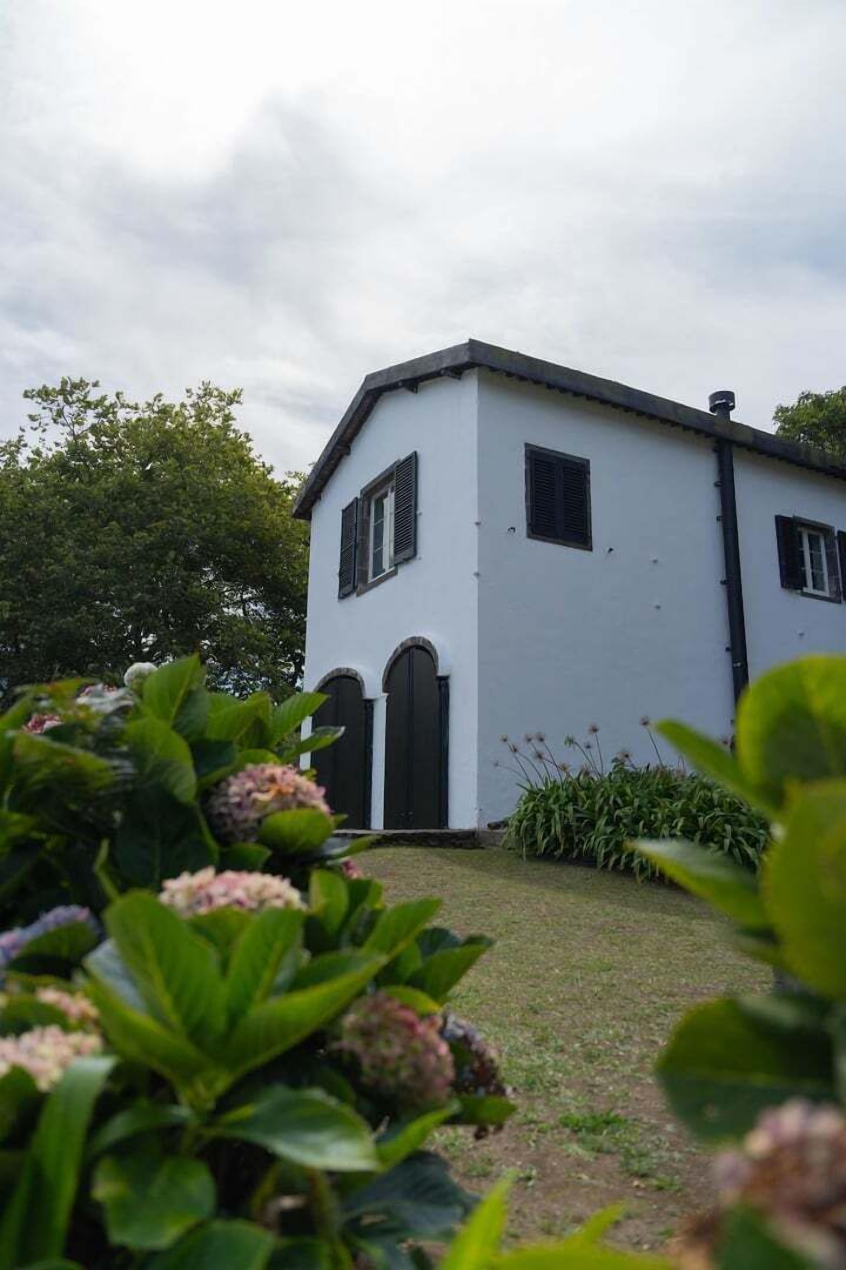 A white building with a gabled roof and black shutters, surrounded by greenery and a cloudy sky.
