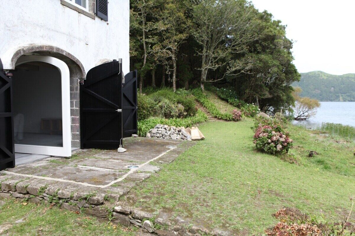 A building with a stone pathway leading to a black gate, surrounded by lush greenery and a lake view.
