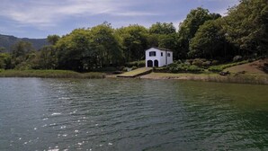 A white building with a small porch by a lake, surrounded by trees and hills.