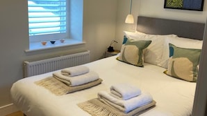 Ein ordentlich bezogenes Bett mit Handtüchern, ein Fenster mit Jalousien, ein Heizkörper und ein an der Wand befestigtes abstraktes Kunstwerk.