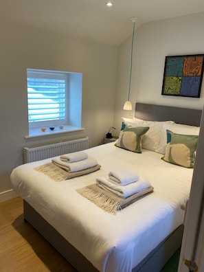Cama bem arrumada com toalhas, janela com persianas, radiador e uma obra de arte abstrata na parede.