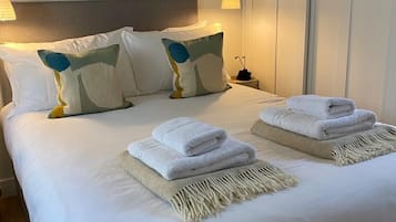 A neatly made bed with white linens, two folded towels, and a decorative pillow.