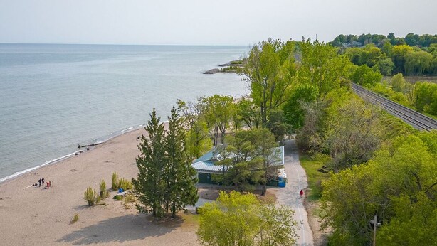 A beach with a path leading to a building with a blue roof, surrounded by greenery and a railway track. - home away home  (Toronto)