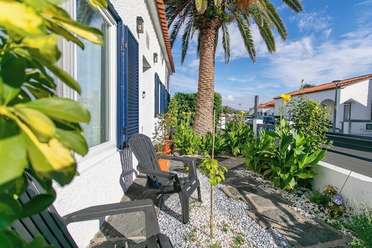 A building with a white exterior and blue shutters, a palm tree, and outdoor seating with a small table and chairs.