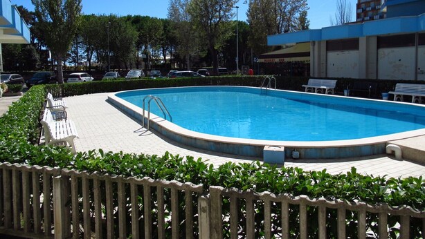 A swimming pool with a wooden deck, surrounded by greenery and benches. - Modern Studio With Shared Pool (Caorle)
