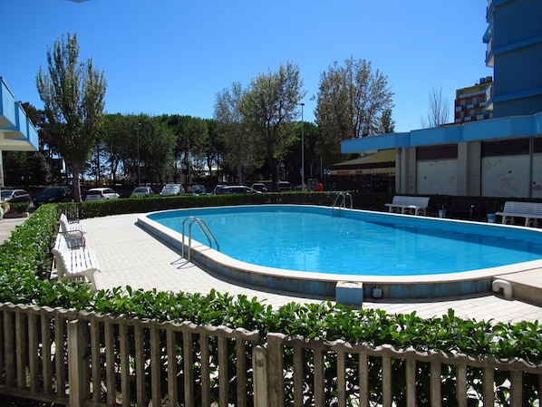 Une piscine dotée d’une terrasse en bois, entourée de verdure et agrémentée de bancs.