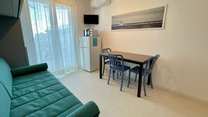 A compact living space with a green sofa, a small dining table with chairs, a refrigerator, a flat-screen TV, and a painting on the wall. - Modern Studio With Shared Pool (Caorle)