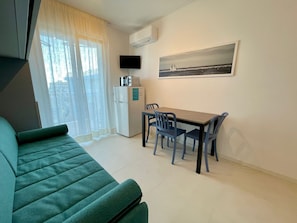 Un espace de vie compact comprenant un canapé vert, une petite table à manger avec des chaises, un réfrigérateur, un téléviseur à écran plat et un tableau accroché au mur.