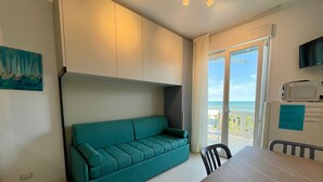 A modern living room with a teal sofa, a dining table, and a view of the ocean through a sliding glass door. - Modern Studio With Shared Pool (Caorle)