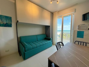 Un salon moderne avec un canapé de couleur turquoise, une table à manger et une vue sur l’océan, visible à travers une porte-fenêtre coulissante.