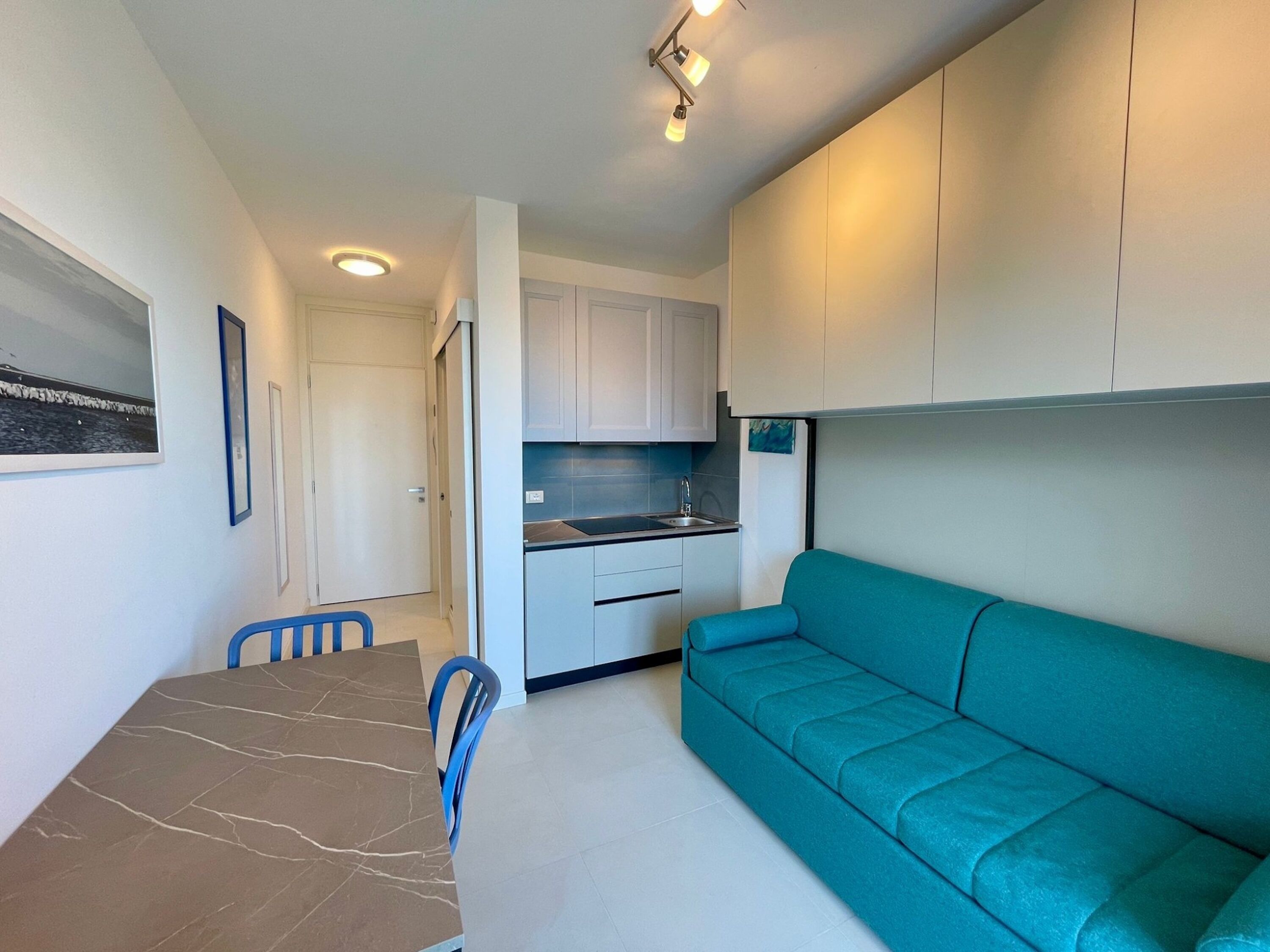 En kompakt opholdsstue med spisebord, blå stole, en turkis sofa, et tekøkken og en dør til et andet rum.