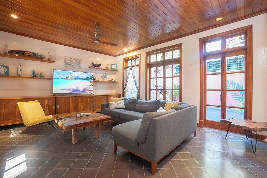 Un salon avec un plafond en bois, un canapé gris, un fauteuil jaune, une table basse en bois et un téléviseur posé sur une console en bois.