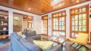 Ein modernes Wohnzimmer mit einer Eckcouch, einem Couchtisch und einem gelben Sessel. Große Fenster mit Holzrahmen und eine Treppe, die in eine andere Ebene führt.