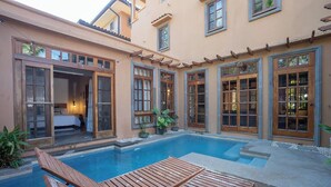 Ein Poolbereich mit Holzliegen, ein Schlafzimmer mit Bett und Nachttisch sowie ein Badezimmer mit Dusche und Toilette.