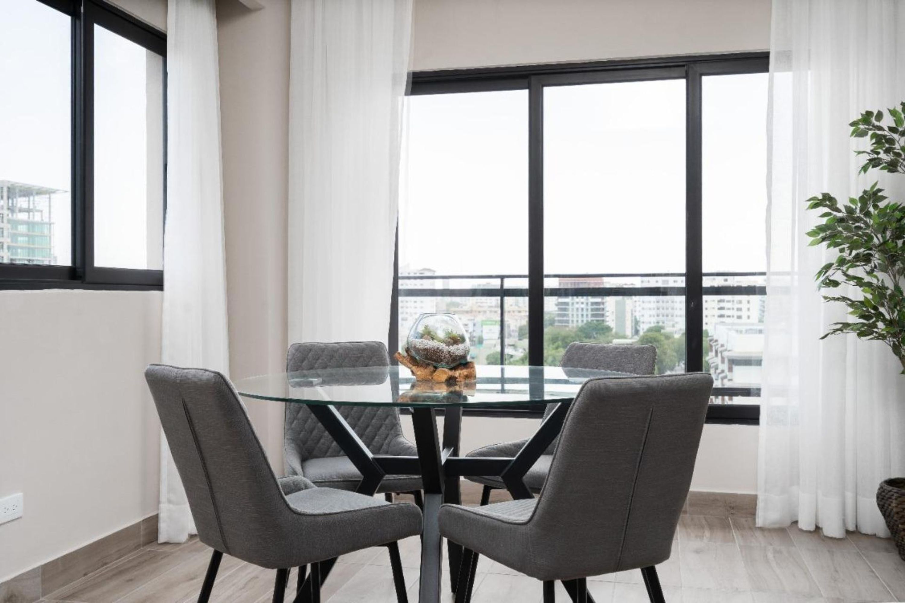 A modern dining area with a glass table and four grey chairs, a decorative globe, and a view of a cityscape through large windows.