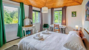 Une chambre d’hôtel avec un plafond en bois, un lit avec du linge de maison blanc, un bureau, une chaise, une petite table et une fenêtre avec des rideaux.