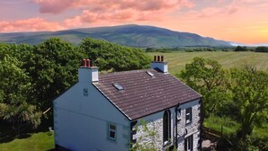 Une maison blanche avec un toit foncé, entourée d’une végétation luxuriante et avec des collines en arrière-plan.