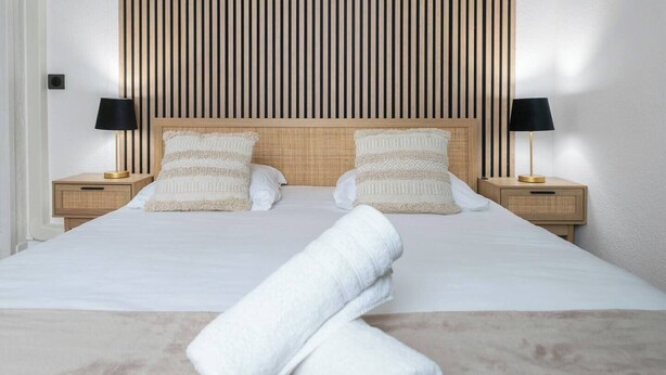 A neatly made bed with white linens, two pillows, and a rolled towel on a wooden headboard. There are two bedside tables with lamps and a wall with vertical wooden slats. - Chez Juliette - 4 rue de Serbie - Vichy (Vichy)