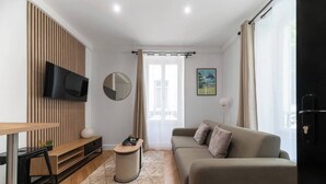 A modern living room with a sofa, a TV mounted on a wooden panel, a small round table, and a window with curtains. - Chez Juliette - 4 rue de Serbie - Vichy (Vichy)