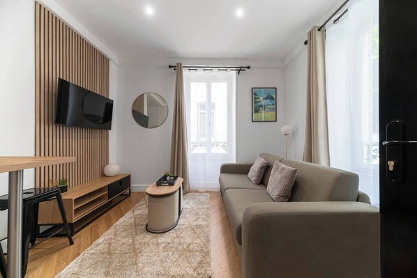 A modern living room with a sofa, a TV mounted on a wooden panel, a small round table, and a window with curtains.