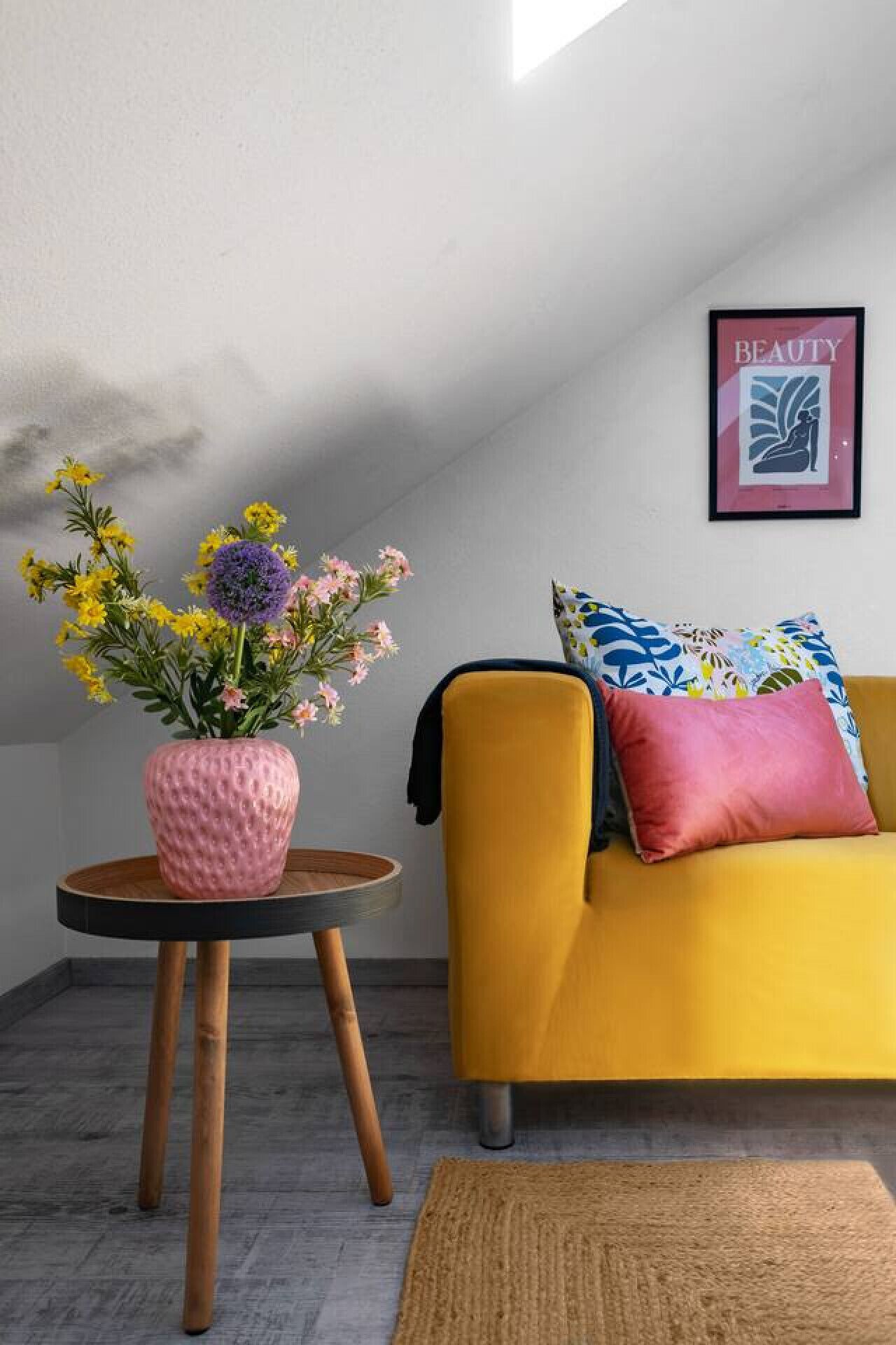 A room with a yellow sofa, a pink textured vase with flowers, and a framed artwork on the wall.