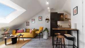 Ein modernes Loft-Apartment mit hohen Decken, einem Essbereich mit Hockern, einer Couch mit bunten Kissen und einer Küche mit dunklen Schränken.