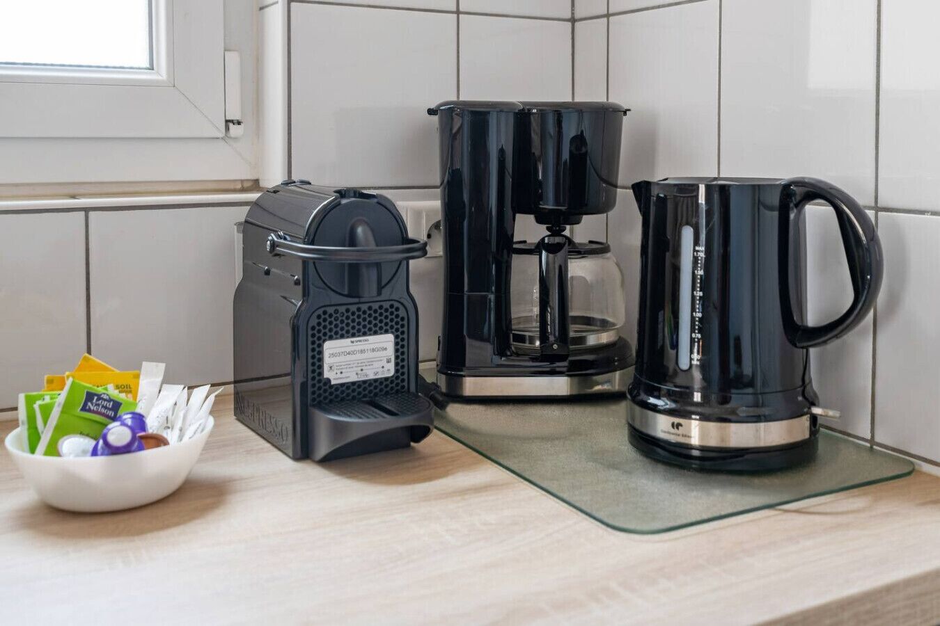 Eine Küche mit einer Kaffeemaschine, einem Wasserkocher und einer Schale mit Kaffeekapseln auf der Arbeitsfläche.