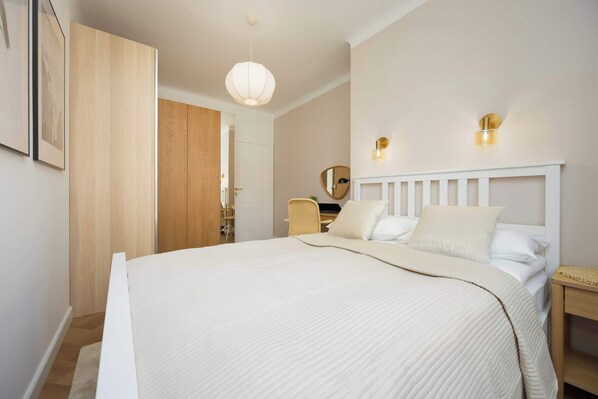 A bedroom with a large bed, a wooden wardrobe, a wicker chair, and a bedside table.