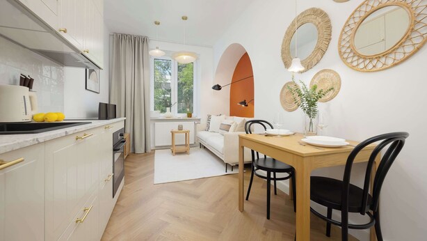 A modern kitchen with white cabinets, a dining area with a wooden table and black chairs, and a living area with a sofa and a large window. - Bright Apartment | Place to Work | Close to the Royal Baths (Warszawa)