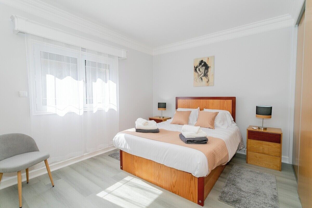 Une chambre à coucher bien aménagée, avec un grand lit, des tables de chevet, une chaise et une fenêtre ornée de rideaux légers.
