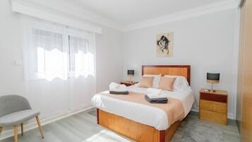 Une chambre à coucher bien aménagée, avec un grand lit, des tables de chevet, une chaise et une fenêtre ornée de rideaux légers.