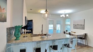 A modern kitchen with a granite countertop, white bar stools, and a dining area with a view of the ocean. - Sea You Soon by Miss Kitty’s (Rockport)