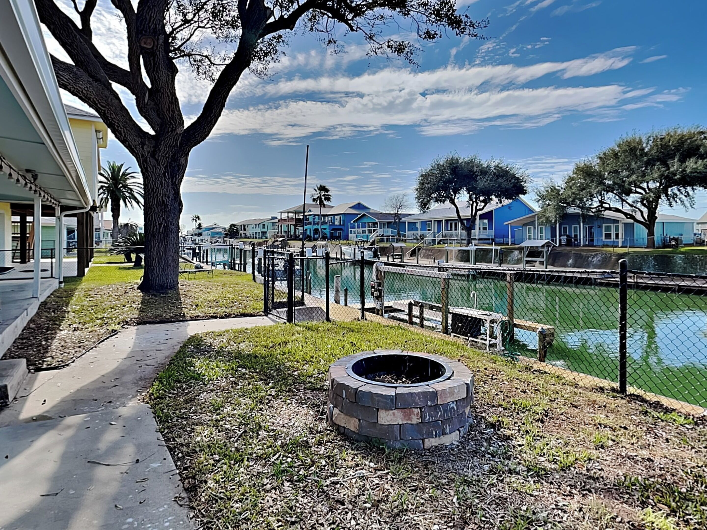 A waterfront property with a fenced pool area, a fire pit, and a pathway leading to a building.
