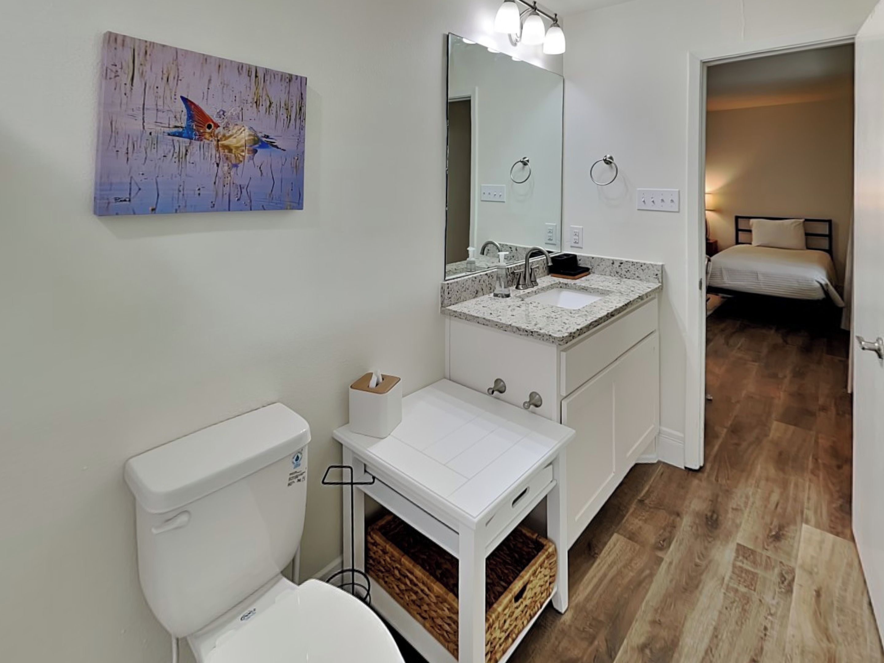 A modern bathroom with a toilet, sink, and mirror. There is a painting on the wall and a bed in the adjacent room.