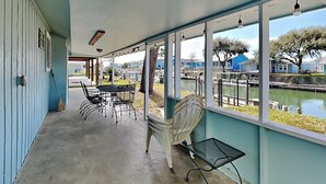 Ein überdachter Außenbereich mit Tisch und Stühlen, einer Bank und Blick auf ein Gewässer sowie Gebäude im Hintergrund.